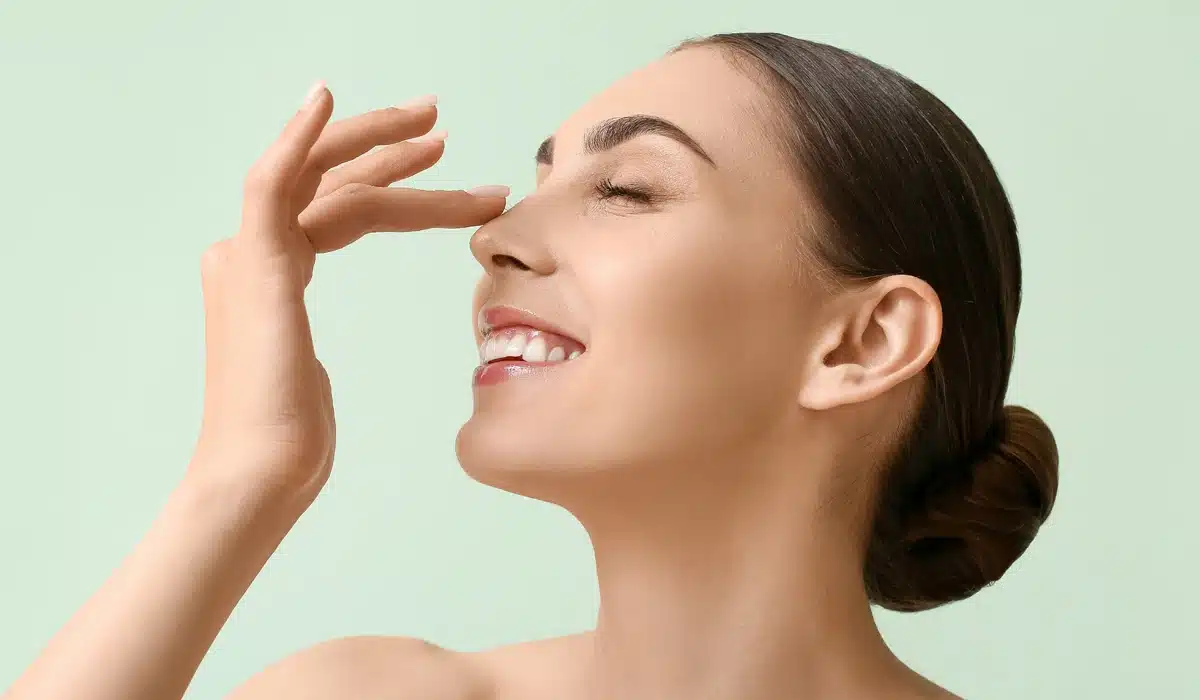 Profile view of a smiling woman with clear skin gently touching her nose, illustrating interest in nasal surgery or rhinoplasty, against a light green background. Ideal for promoting cosmetic or reconstructive nasal procedures